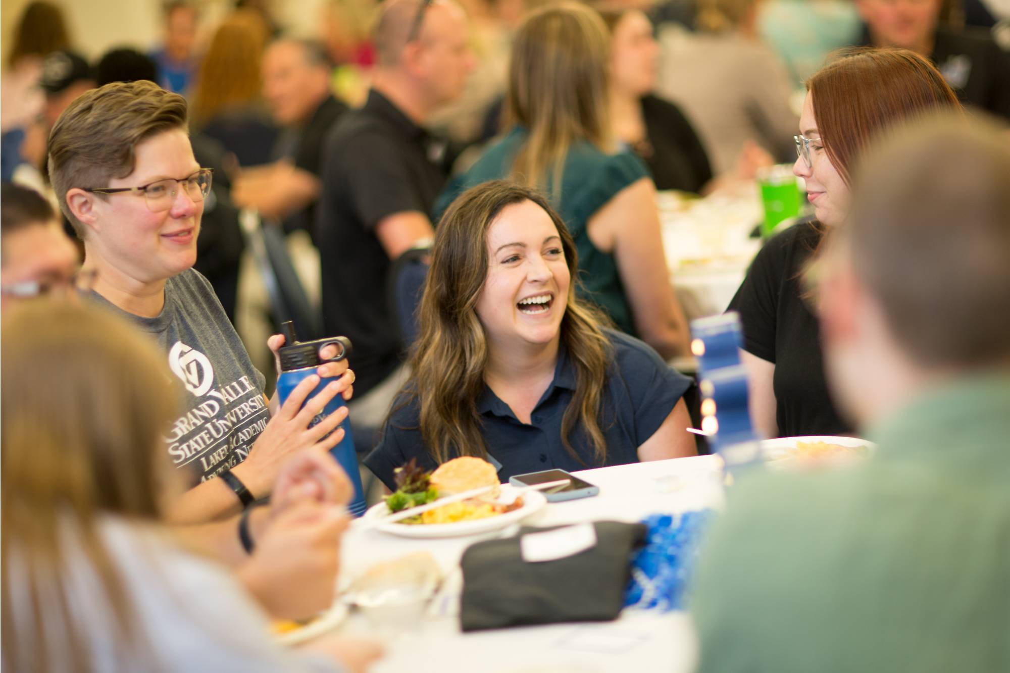 A group of women laughing together around a table. They are eating lunch.